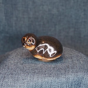 May include: A small, smooth ceramic figurine of a seal with a glossy dark brown glaze. The seal has a rounded body, a small head with visible eyes and a snout, and a lighter tan-colored base. The surface reflects light, highlighting the curves.