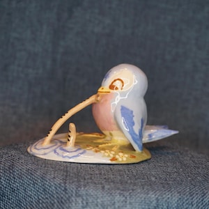 May include: A ceramic figurine of a small bird with light blue and pink plumage, perched on a base decorated with painted flowers. The bird has a long, thin, curved beak that extends downward toward the base, which features blue water ripples.