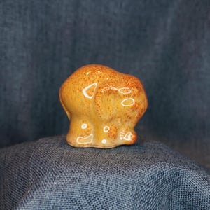 May include: A small ceramic elephant figurine in a yellow and orange glaze. The elephant is standing with its trunk curled up.