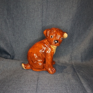 May include: A brown ceramic figurine of a puppy sitting with its head tilted to the side. The puppy has a brown and white coat and black eyes.