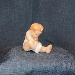 May include: A ceramic figurine of a baby in a seated position, with a light skin tone and light brown hair. The baby is wearing a white diaper with blue trim and a light blue top. The figurine is on a blue denim surface.