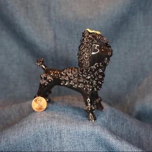 May include: A black ceramic poodle figurine with a textured coat and a small yellow accent on its head. The poodle is standing on a blue surface, with a penny placed near its front legs for size comparison. The figurine has a glossy finish.