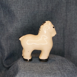 May include: A small, cream-colored ceramic horse figurine with a glossy finish. The horse features dark brown painted details on its mane, tail, eyes, and hooves. The design is simple and stylized, showing the animal in a standing profile position.