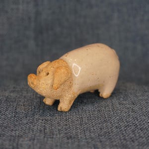 May include: A ceramic pig figurine with a light beige body and tan head and ears. The small, handcrafted pig has a glossy finish and is posed in a standing position. This decorative item could be used as a home decor accent.