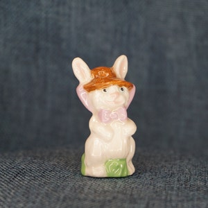 May include: A white ceramic figurine of a rabbit wearing a brown hat with a pink bow tie. The rabbit is sitting on a green base.
