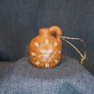 May include: A small, orange ceramic jug with a handle and a decorative bird design. The bird is white with a green and pink accent. A natural twine loop is attached to the handle. The jug is sitting on a blue surface.