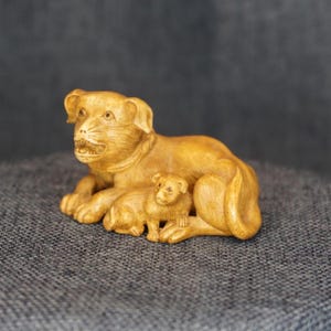 May include: A detailed wooden carving of a mother dog and her puppy. The mother dog is lying down, and the puppy is sitting in front of her. The carving is a light brown color and has intricate details.