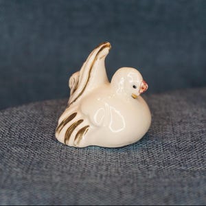 May include: A small, cream-colored ceramic dove figurine with gold accents on the wings and tail. The dove has a red beak and is perched on a surface. The figurine is a decorative item.