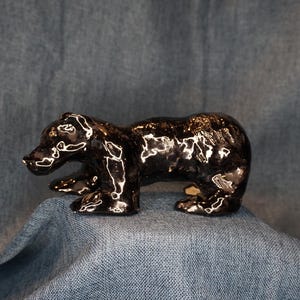 May include: A small, glossy, dark brown ceramic bear figurine standing in a quadrupedal position. The surface of the bear has a reflective, high-shine glaze that catches the light, highlighting the sculpted contours of its head, body, and legs.