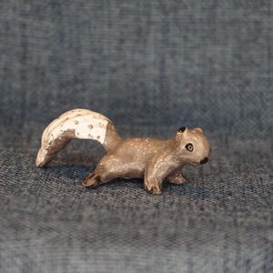 May include: A small, gray squirrel figurine with a white tail and black eyes. The figurine is made of a textured material and has a realistic appearance.