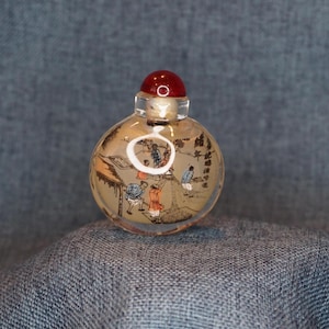 May include: A small, clear glass snuff bottle with a red stopper. The bottle is decorated with a detailed, hand-painted scene of people working. The stopper is a deep red color, and the bottle has a cork closure.