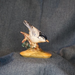 May include: A ceramic figurine of a small bird with a black head, white chest, and grey-toned wings, perched on a textured brown branch. The bird has a small orange beak and is positioned facing right, resting on a smooth, rounded base.