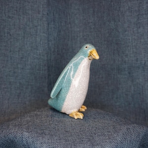 May include: A ceramic penguin figurine with a crackle glaze finish. The penguin has a light blue body, white belly, and yellow beak and feet. The figurine is standing on a blue surface against a blue background.