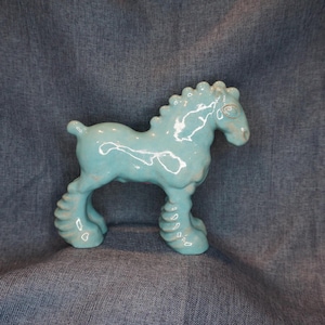 May include: A turquoise ceramic horse figurine with a glossy finish. The horse has a stylized design with rounded features, including a textured mane and thick legs. The figurine is standing on a blue background.