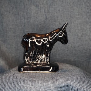 May include: A small, glossy, dark brown ceramic figurine of a donkey standing on a rectangular base. The surface of the donkey is highly reflective, showing bright white highlights from the light source, with a smooth, curved silhouette.
