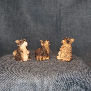 May include: Three small raccoon figurines in various poses. The figurines are primarily brown and tan, with black markings around the eyes and on the tail. They are set against a textured, blue-gray background.