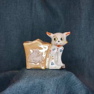 May include: A white ceramic cat figurine with a brown and orange house on its back. The cat has large, round eyes and a spotted pattern. The house has a small, purple flower design.