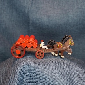 May include: A miniature wooden toy depicting a horse-drawn cart carrying red logs. The cart has red wheels and a small figure of a person. The horse is brown with black mane and tail.