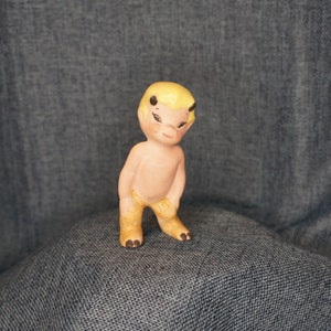 Puede incluir: Pequeña figura de cerámica de un querubín desnudo y sonriente con pelo rubio y pequeños cuernos negros. El querubín tiene un tono de piel rosa claro y pies y piernas de color canela. La figura está sobre una superficie de mezclilla azul.