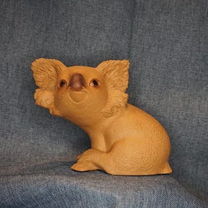 May include: A light brown koala figurine with textured fur detailing, large rounded ears, a dark brown nose, and dark eyes. The koala is depicted in a seated position with its body angled slightly to the left, showcasing a smooth, matte finish.