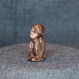 May include: A small, brown ceramic monkey figurine in a seated position. The monkey has a light-colored face and is detailed with dark brown accents. The figurine is displayed on a textured, blue-gray surface.