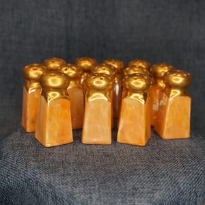 May include: A collection of small salt and pepper shakers featuring a square base with a marbled orange finish and a rounded, metallic gold top. Each shaker has a series of small holes on the top surface for dispensing seasoning.