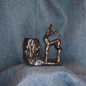 May include: A black ceramic deer figurine standing on a tree stump. The deer is in a standing position with its head turned to the side. The tree stump is textured to look like bark.