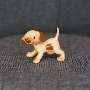 May include: A small ceramic puppy figurine in a standing pose. The puppy has a cream-colored body with brown spots and ears. Its eyes and nose are dark, and it has a glossy finish.