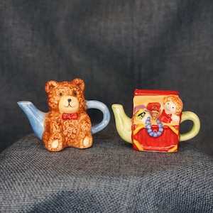 May include: Two decorative teapots. One is shaped like a brown teddy bear with a red bow tie and blue handle and spout. The other is a red teapot with a yellow and green handle and spout, and a design of a girl.