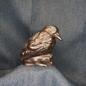 May include: A bronze-colored bird figurine perched on a small base. The bird has detailed feather textures and a slightly weathered appearance. The sculpture is a decorative item, likely for home decor.