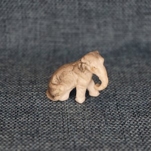 May include: A small, white ceramic elephant figurine with gray detailing. The elephant has its trunk curled up and its eyes are painted black.