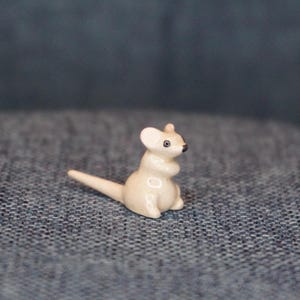 May include: A small, off-white ceramic kangaroo figurine. The kangaroo is sitting upright with its tail extended. It has black eyes and a small, rounded ear. The figurine is on a textured, blue surface.