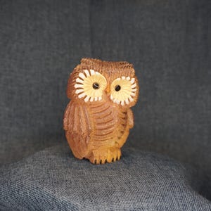 May include: A small, hand-carved wooden owl figurine featuring a light brown finish with detailed feather textures. The owl has two prominent, circular, cream-colored facial discs surrounding dark, round eyes and a small, pointed beak.
