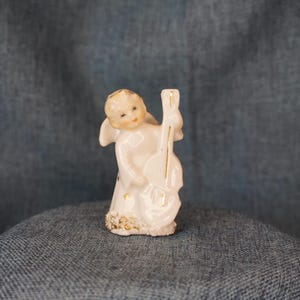 May include: A small, white ceramic angel figurine playing a cello. The angel has delicate features, small wings, and gold accents on the cello and the hem of its robe. The figurine is standing on a decorative base.