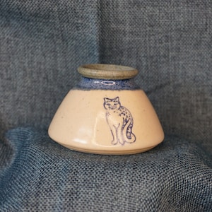 May include: A small, cream-colored ceramic vase with a blue glaze around the rim. The vase has a stylized image of a cat with blue spots on a white background.