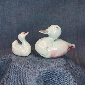 May include: Two ceramic duck figurines in a glossy finish. The larger duck is positioned on the right, featuring a pale blue body with soft pink accents on the chest and wing. The smaller duck on the left mirrors this color scheme and design.