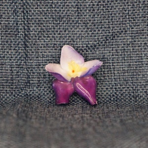 May include: A purple and white ceramic flower with a yellow center. The flower is a decorative piece and has a three-dimensional design.