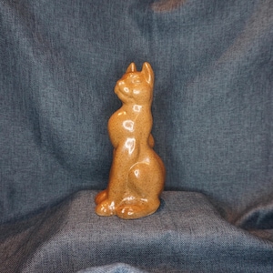 May include: A ceramic cat figurine in a seated position, with a glossy, speckled brown finish. The cat has pointed ears and a curved tail, showcasing a detailed, handcrafted appearance. The figurine is a decorative item.