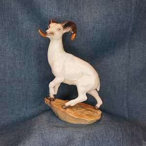 May include: A ceramic figurine of a mountain goat with a cream-colored body and dark brown curved horns. The goat is depicted in a standing position with one front leg raised, perched upon a textured, light brown rock-like base.
