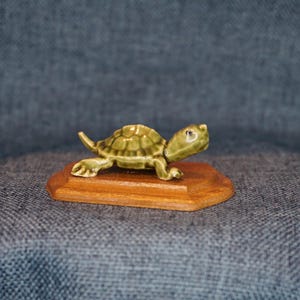 May include: A small green ceramic turtle figurine walking on a wooden base. The turtle has a detailed shell and realistic features.
