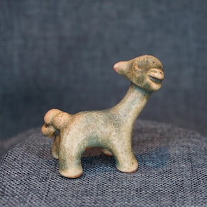 May include: A small, hand-sculpted ceramic figurine of a llama with a textured, mottled surface in shades of muted green, beige, and brown. The figure features a long neck, a small head with pointed ears, and a short, rounded tail, standing on four legs.