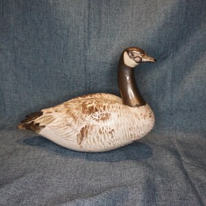 May include: A ceramic goose figurine with a brown head and neck, white body, and brown and white speckled markings. The goose is in a resting position with its head turned to the side.