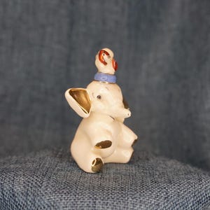 May include: A white ceramic elephant figurine with gold accents. The elephant is wearing a blue and red hat with a gold band.