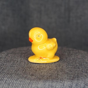 May include: A small, yellow ceramic duck figurine with an orange beak and white accents. The duck is standing on a small, round base. The figurine is a decorative item.