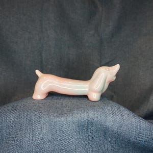May include: A ceramic dachshund figurine in a light pink color with subtle green and orange accents. The dog has a long body, short legs, and a raised head, giving it a playful appearance. This decorative item is a charming addition to any home decor.