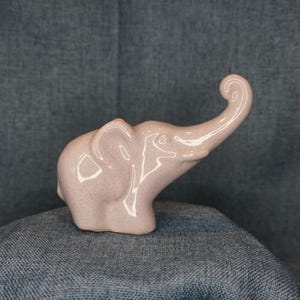 May include: A ceramic elephant figurine in a light pink color with a speckled texture. The elephant has its trunk raised and ears extended. The figurine is a decorative item.