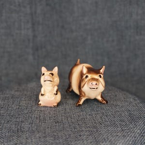 May include: Two ceramic pig figurines. One pig is standing upright, the other is on all fours. The pigs are primarily tan and brown with black accents. The figurines have a glossy finish and are set against a blue-gray background.