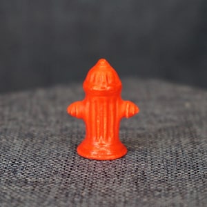 May include: A small, bright orange fire hydrant figurine. The miniature hydrant has a conical top, two side nozzles, and a base. The object is set against a textured gray background.