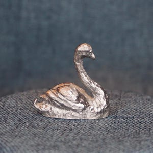 May include: A small, silver-colored swan figurine. The swan is sculpted with textured details, showcasing its feathers and curved neck. The figurine is resting on a textured, blue surface, with a darker blue backdrop.