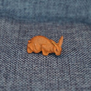 May include: A small, brown, wooden rabbit figurine. The rabbit is lying on its belly with its head turned to the side. The rabbit has long ears and a short tail.